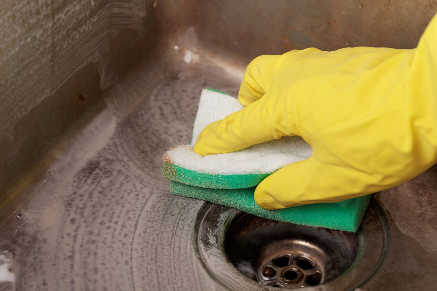 Close-up of hand in yellow rubber glove cleaning an old stainless sink with a mild abrasive