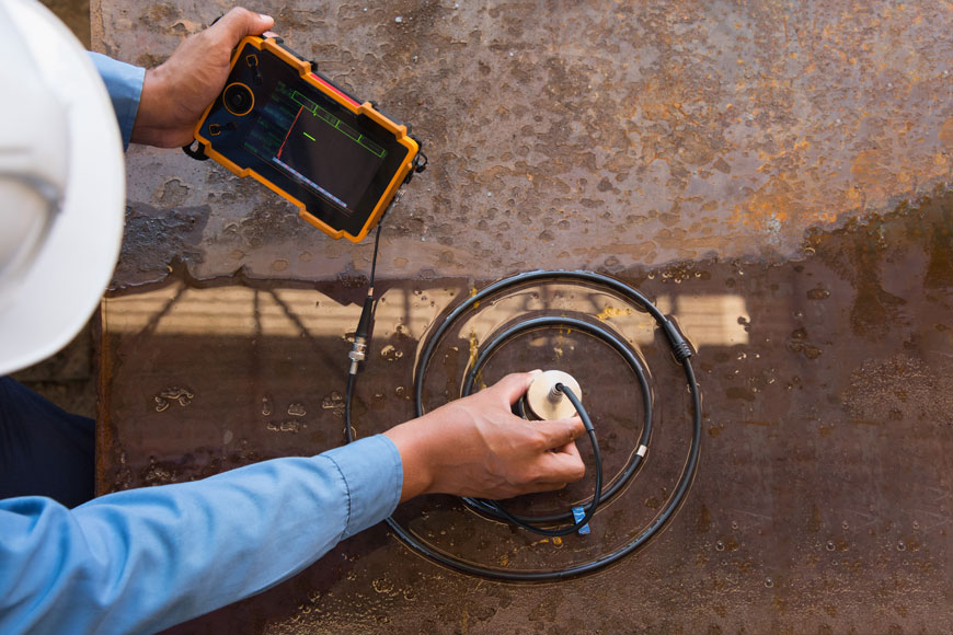 A technician uses a metal probe centered in a spiral of wire to test a wet, rusty metal surface