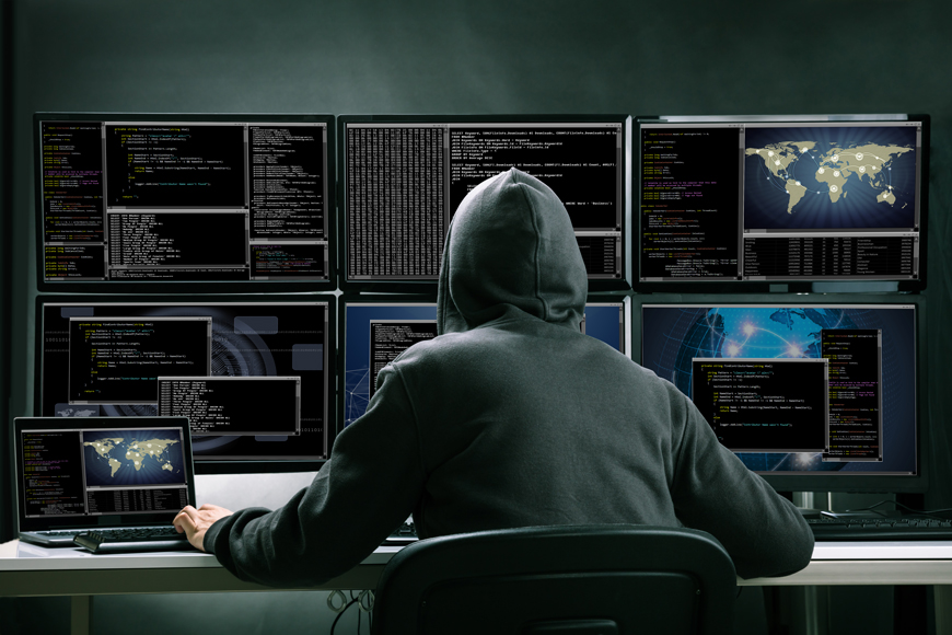 A man in a hoodie sits in front of a bank of computer screens